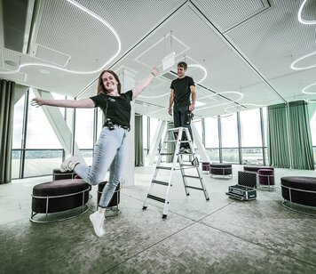 Junge Frau hüpft in die Luf, daneben ein junger Mann auf einer Leiter | © MUNK GmbH
