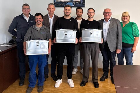 Die Geschäftsleitung und Ausbilder gratulieren  drei Auszubildenen zur bestandenen Abschlussprüfung und überreichen eine Kiste mit kleinen Geschenken an die Absolventen.