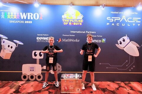 Zwei junge Männer posieren vor einem Bühnenhintergrund mit dem Titel "The Future of Robots" sowie Logos verschiedener Firmen.